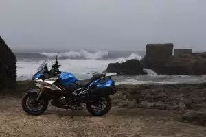 Suzuki-Motorrad steht auf felsiger Küste vor dem Meer mit Wellen und alten Steinmauern im Hintergrund