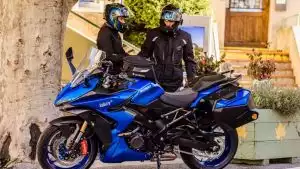 Zwei Motorradfahrer in Schutzkleidung neben einer blauen Suzuki Sporttourer vor einem Baum.