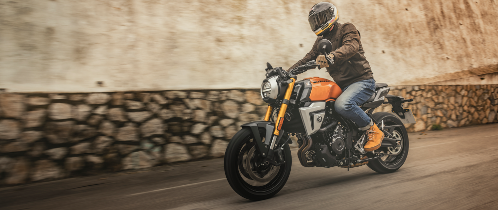 Fahrer auf einer orangefarbenen Suzuki GSX-8T im klassischen Roadster-Look, unterwegs auf einer Straße mit Steinmauer im Hintergrund – urban und stylisch.