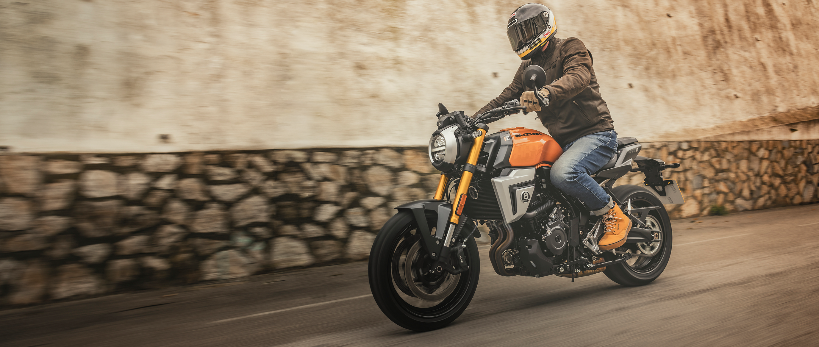 Fahrer auf einer orangefarbenen Suzuki GSX-8T im klassischen Roadster-Look, unterwegs auf einer Straße mit Steinmauer im Hintergrund – urban und stylisch.