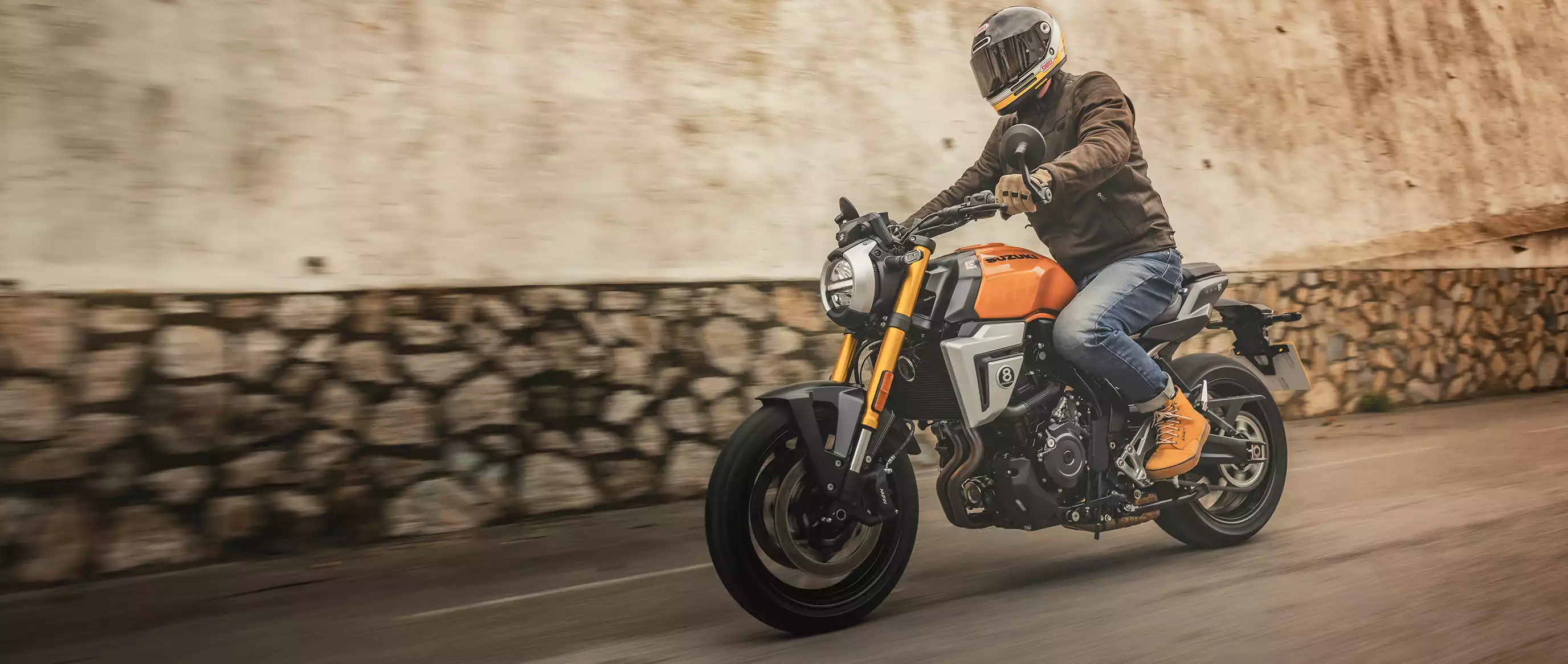 Fahrer auf einer orangefarbenen Suzuki GSX-8T im klassischen Roadster-Look, unterwegs auf einer Straße mit Steinmauer im Hintergrund – urban und stylisch.