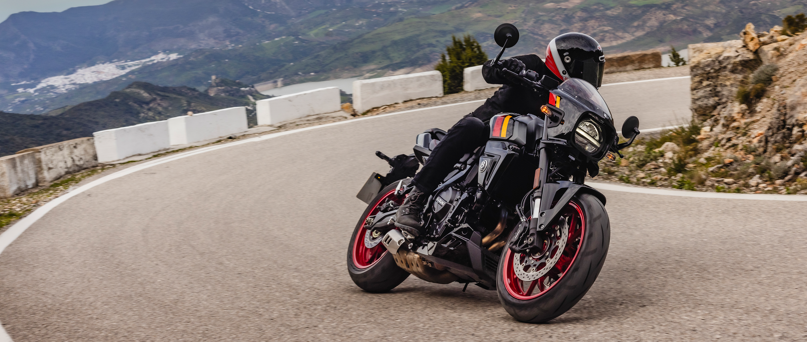 Fahrer auf einer schwarzen Suzuki GSX-8TT mit roten Felgen und Café-Racer-Elementen, sportlich in Schräglage auf einer kurvigen Bergstraße – vor beeindruckender Landschaftskulisse.