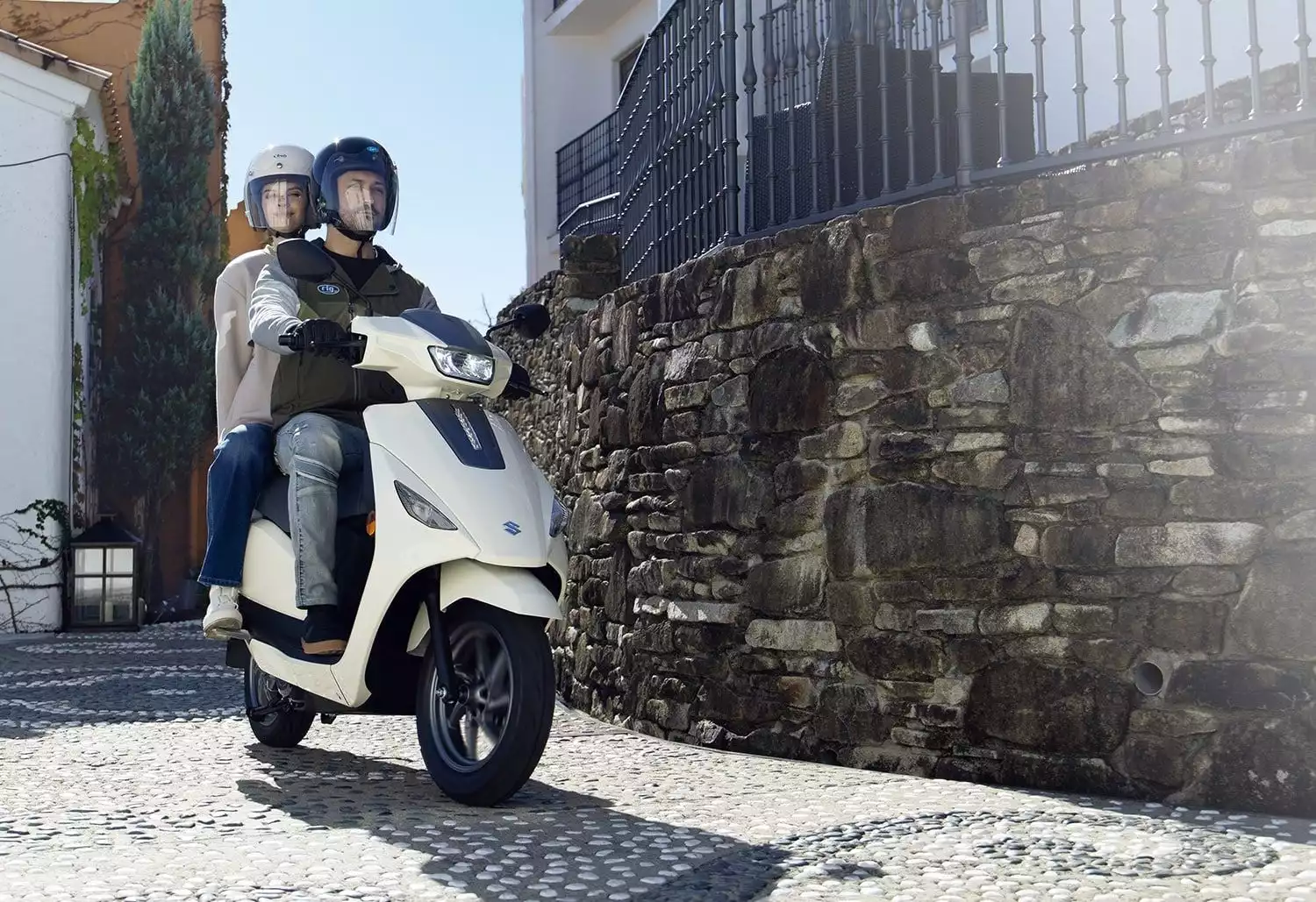 Zwei Personen fahren auf einem weißen Suzuki e-Address E-Scooter durch eine sonnige, enge Gasse mit Kopfsteinpflaster und alten Gebäuden.