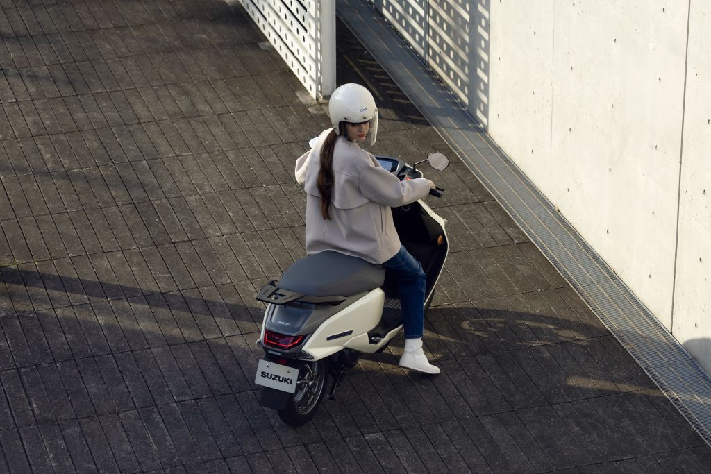 Person mit weißem Helm sitzt auf einem weißen Suzuki-Roller und blickt über die Schulter, umgeben von Beton- und Metallstrukturen.