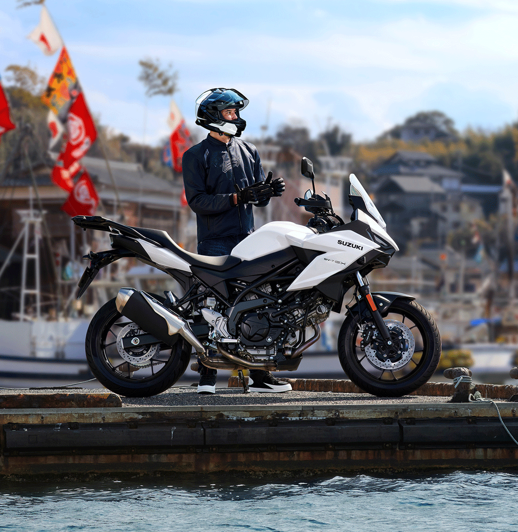 Ein Motorradfahrer steht am Hafen neben einer weißen Suzuki SV-7GX und zieht seine Handschuhe an; im Hintergrund sind Boote und bunte Fahnen zu sehen.
