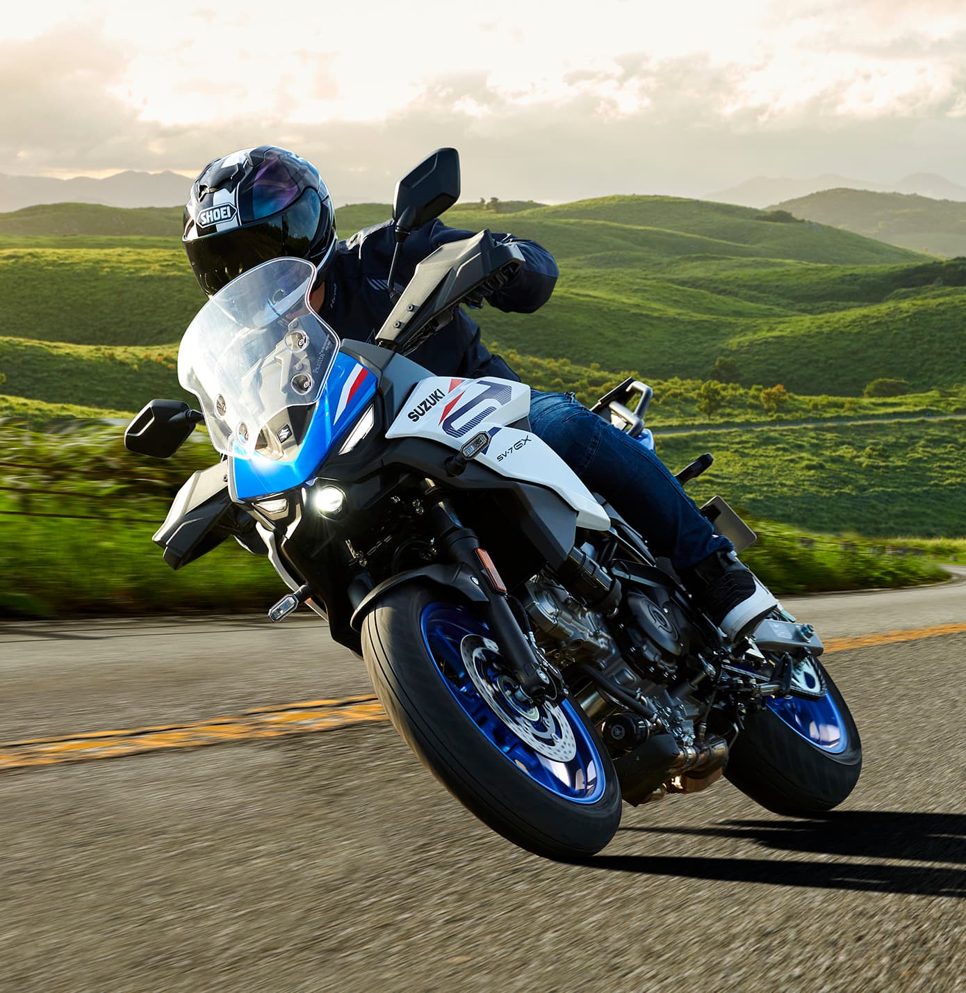 Ein Motorradfahrer fährt eine blau-weiße Suzuki SV-7GX in tiefer Schräglage durch eine kurvige Landstraße; grüne Hügel und warmes Abendlicht im Hintergrund.