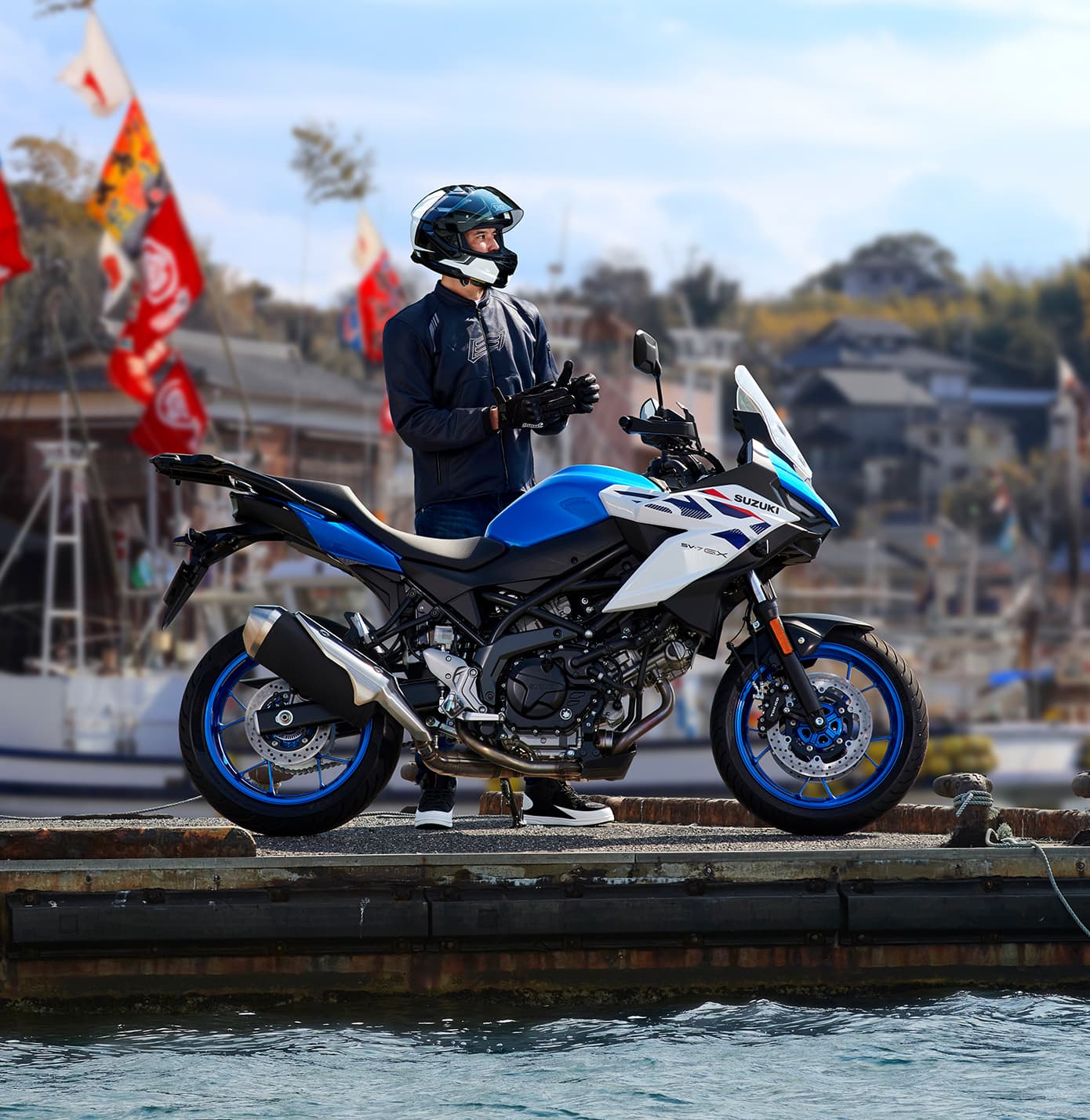 Ein Motorradfahrer steht am Hafen neben einer blau-weißen Suzuki SV-7GX und zieht seine Handschuhe an; im Hintergrund sind Boote und bunte Fahnen zu sehen.