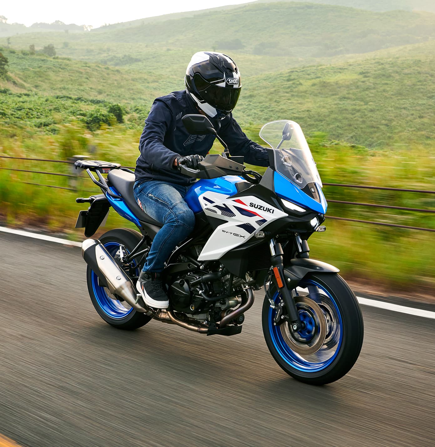 Ein Motorradfahrer fährt eine blau-weiße Suzuki SV-7GX auf einer Landstraße, umgeben von grünen Hügeln; der Fahrer trägt dunkle Schutzkleidung und Helm.