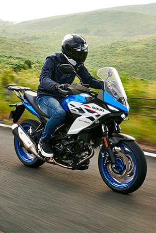 Ein Motorradfahrer fährt eine blau-weiße Suzuki SV-7GX auf einer Landstraße, umgeben von grünen Hügeln; der Fahrer trägt dunkle Schutzkleidung und Helm.