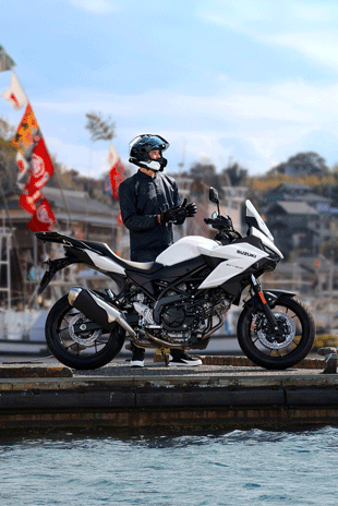 Ein Motorradfahrer steht am Hafen neben einer weißen Suzuki SV-7GX und zieht seine Handschuhe an; im Hintergrund sind Boote und bunte Fahnen zu sehen.