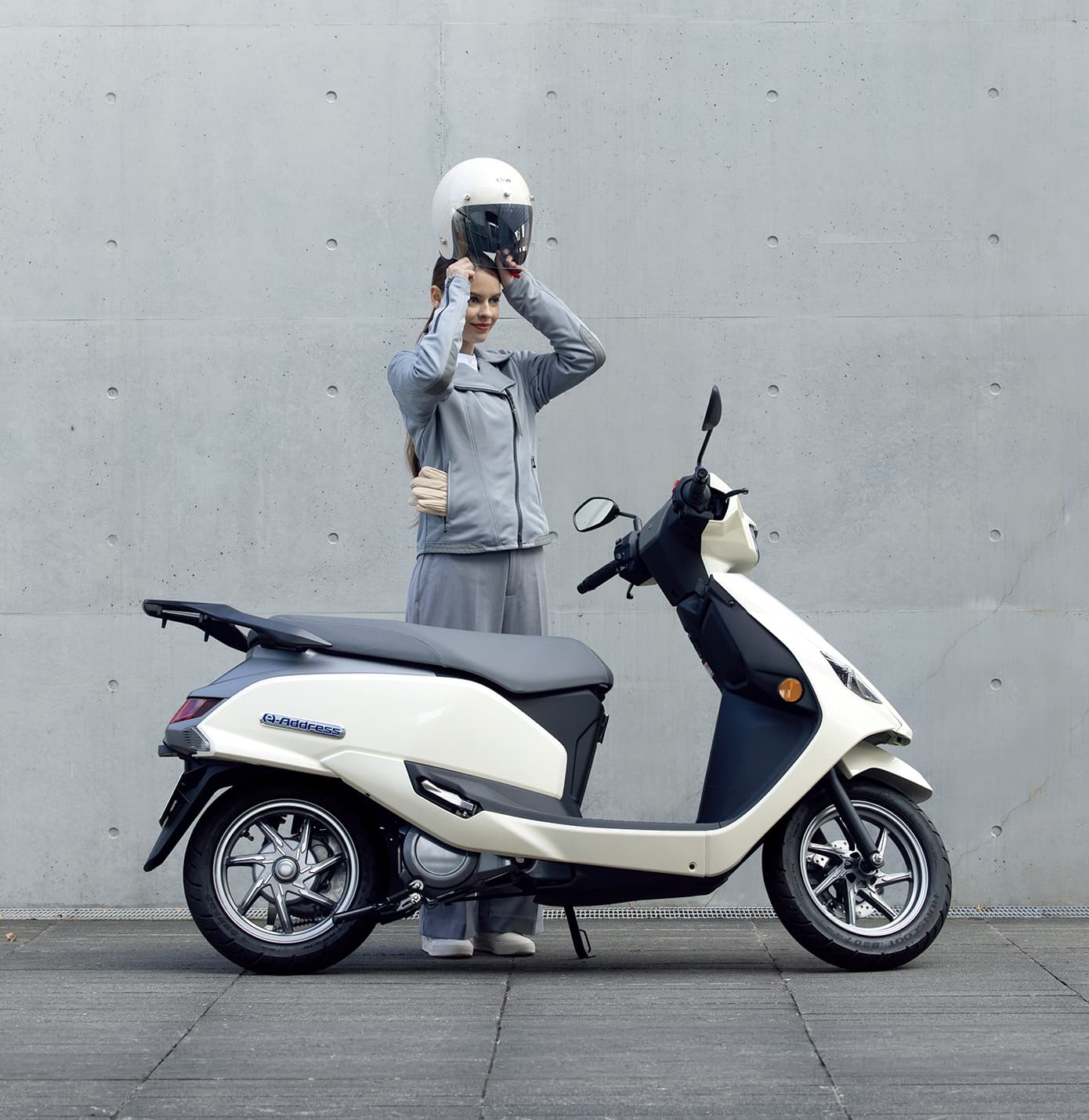 Eine Person stellt einen weißen Helm auf den Kopf, während sie neben einem weißen Suzuki-e-Address-Elektroroller steht; Hintergrund ist eine schlichte Betonwand.