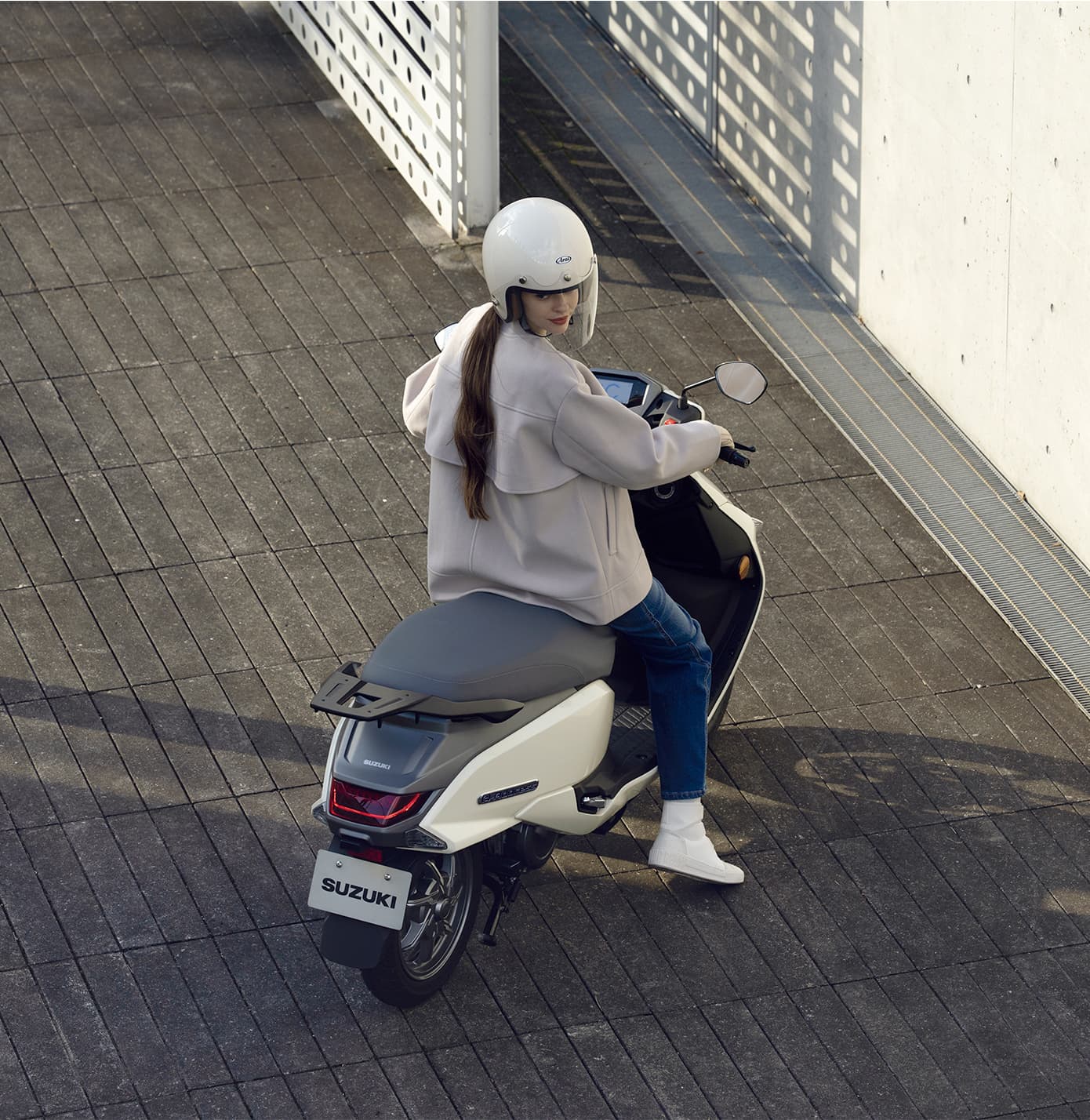 Eine Person sitzt mit Helm auf einem weißen Suzuki-e-Address-Elektroroller und blickt über die Schulter zurück; das Bild ist aus erhöhter Perspektive aufgenommen.