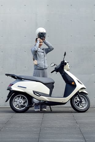 Eine Person stellt einen weißen Helm auf den Kopf, während sie neben einem weißen Suzuki-e-Address-Elektroroller steht; Hintergrund ist eine schlichte Betonwand.