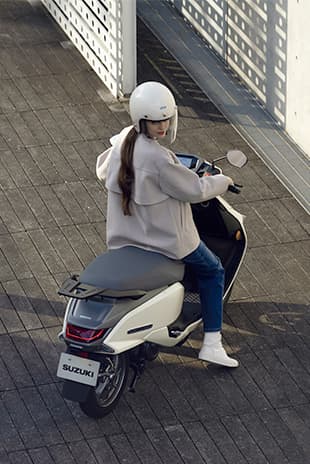 Eine Person sitzt mit Helm auf einem weißen Suzuki-e-Address-Elektroroller und blickt über die Schulter zurück; das Bild ist aus erhöhter Perspektive aufgenommen.
