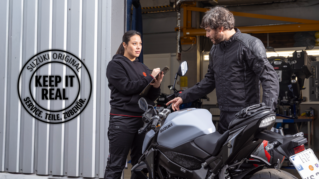 Suzuki Service-Mitarbeiterin im Gespräch mit einem Kunden an seiner grauen Suzuki GSX-S950 vor der Werkstatt. Links im Bild das runde Logo „Suzuki Original – Keep it real. Service. Teile. Zubehör.“ als Hinweis auf originalgetreuen Service und Ersatzteile.