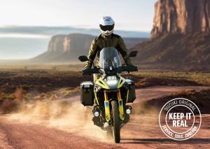 Das Bild zeigt ein Motorrad, das auf einer staubigen Straße in einer landschaftlich reizvollen Umgebung fährt. Das Motorrad ist mit Seitenkoffern ausgestattet und verfügt über zahlreiche weitere Zubehörteile, die einen robusten und abenteuerlichen Charakter unterstreichen. Im rechten unteren Bereich befindet sich ein rundes Logo mit dem Schriftzug „SUZUKI ORIGINAL – KEEP IT REAL – SERVICE. TEILE. ZUBEHÖR.“ Die Szene vermittelt Freiheit, Abenteuer und Authentizität.