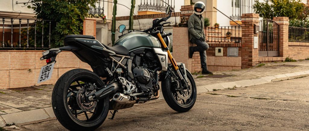 Eine Suzuki GSX-8T in mattem Grün-Schwarz steht auf einer Wohnstraße, im Hintergrund lehnt ein Motorradfahrer mit Helm lässig an einer Mauer vor einem Haus.