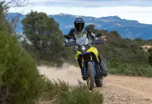 Eine Person fährt eine gelbe Suzuki V-Strom auf einem staubigen Schotterweg durch eine landschaftlich schöne Umgebung mit grünen Büschen und Bergen im Hintergrund. Das Motorrad wirbelt Staub auf und vermittelt Dynamik sowie Abenteuerlust im Gelände.