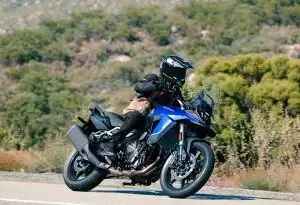 Eine Person fährt eine blaue Suzuki V-Strom 800DE auf einer kurvigen Straße durch eine landschaftlich grüne Umgebung. Das Motorrad ist in sportlicher Schräglage, die Szene vermittelt Dynamik, Fahrspaß und Abenteuer.