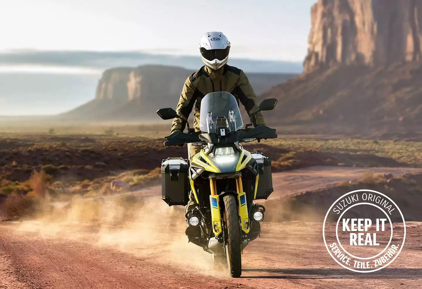 Das Bild zeigt ein Motorrad, das auf einer staubigen Straße in einer landschaftlich reizvollen Umgebung fährt. Das Motorrad ist mit Seitenkoffern ausgestattet und verfügt über zahlreiche weitere Zubehörteile, die einen robusten und abenteuerlichen Charakter unterstreichen. Im rechten unteren Bereich befindet sich ein rundes Logo mit dem Schriftzug „SUZUKI ORIGINAL – KEEP IT REAL – SERVICE. TEILE. ZUBEHÖR.“ Die Szene vermittelt Freiheit, Abenteuer und Authentizität.