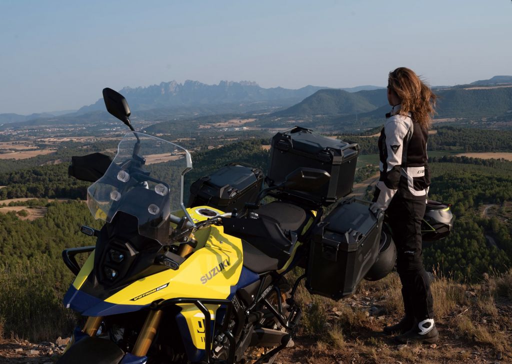 Eine Motorradfahrerin steht neben einem gelben Suzuki-Motorrad mit großen schwarzen Seitenkoffern auf einem Hügel und blickt in die Ferne. Im Hintergrund ist eine malerische Landschaft mit Bergen und Tälern zu sehen.
