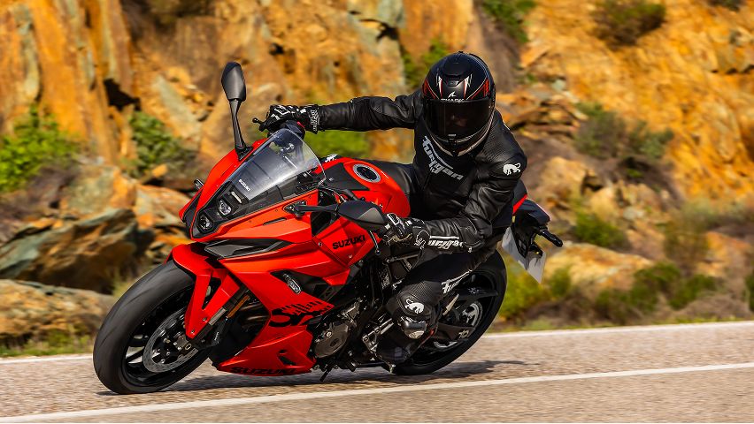 Sportliches blaues Suzuki-Motorrad in voller Fahrt auf einer Rennstrecke; die fahrende Person trägt vollständige Schutzkleidung und beugt sich in dynamischer Haltung über den Lenker. Der Hintergrund ist durch die Bewegung verwischt.