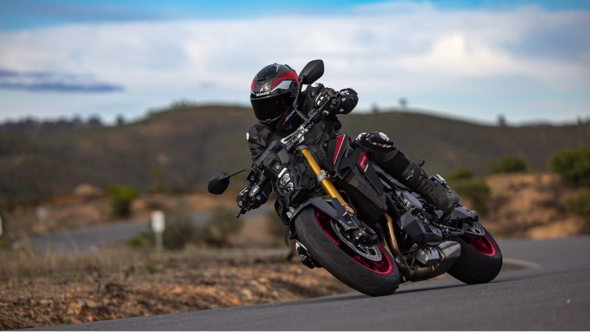 Motorrad in dynamischer Schräglage auf einer kurvigen Landstraße; die fahrende Person trägt dunkle Schutzkleidung und Helm. Im Hintergrund liegen hügelige Landschaft und ein teils bewölkter Himmel.