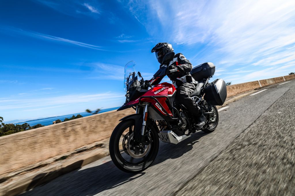 Rotes Suzuki‑Adventure‑Motorrad mit Seitentaschen und Topcase fährt auf einer sonnigen Küstenstraße; die fahrende Person trägt komplette Schutzkleidung und Helm. Im Hintergrund sind Meer, Himmel und leichte Bewölkung zu sehen.