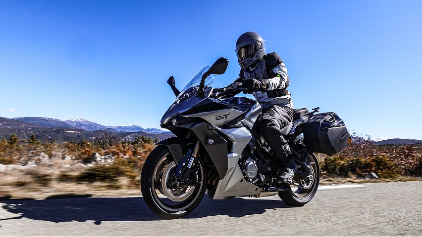 Graues Suzuki‑Touring‑Motorrad mit Seitentaschen fährt über eine sonnige Landstraße; die fahrende Person trägt vollständige Schutzkleidung und Helm. Im Hintergrund erstrecken sich Berge und herbstliche Landschaft unter klarem blauem Himmel.