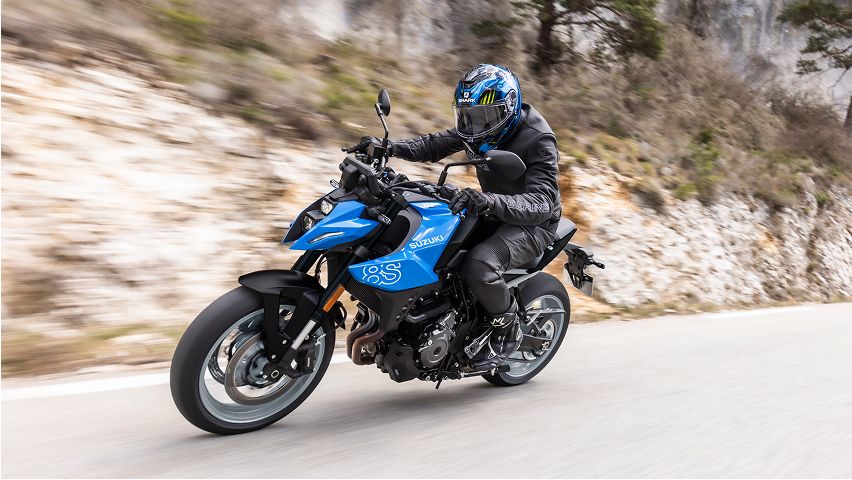 Blaues Suzuki‑Motorrad fährt dynamisch durch eine kurvige Straße entlang einer Felswand; die fahrende Person trägt dunkle Schutzkleidung und Helm. Der Hintergrund ist leicht verwischt durch die Bewegung.