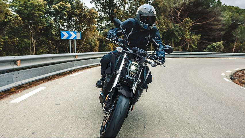 Motorradfahrer in dunkler Schutzkleidung fährt ein schwarzes Naked‑Bike dynamisch durch eine enge Rechtskurve; im Hintergrund stehen dichte Bäume und ein blau‑weißes Richtungsschild.