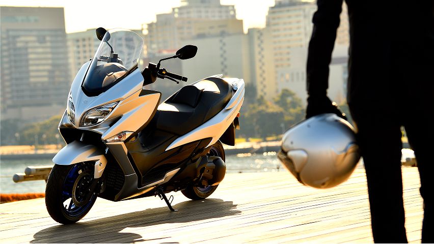 Silberner Maxi‑Scooter steht auf einem Holzsteg am Wasser; im Hintergrund sind hohe Gebäude zu sehen. Rechts im Vordergrund hält eine Person in dunkler Kleidung einen Motorradhelm in der Hand.