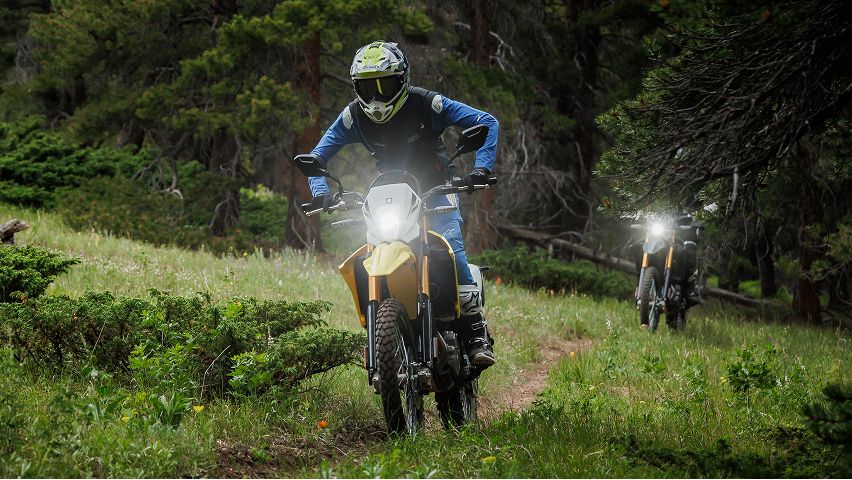 Zwei Offroad-Motorräder fahren auf einem schmalen Waldpfad durch dichtes Grün; die vordere Person trägt blaue Schutzkleidung und Helm, das Scheinwerferlicht ist eingeschaltet. Im Hintergrund folgt ein zweites Motorrad zwischen den Bäumen.