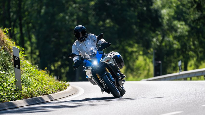 Motorradfahrer in heller Schutzkleidung fährt ein blaues Touring‑Motorrad in Schräglage durch eine sonnige, kurvige Landstraße; dichtes grünes Laubwerk bildet den Hintergrund.