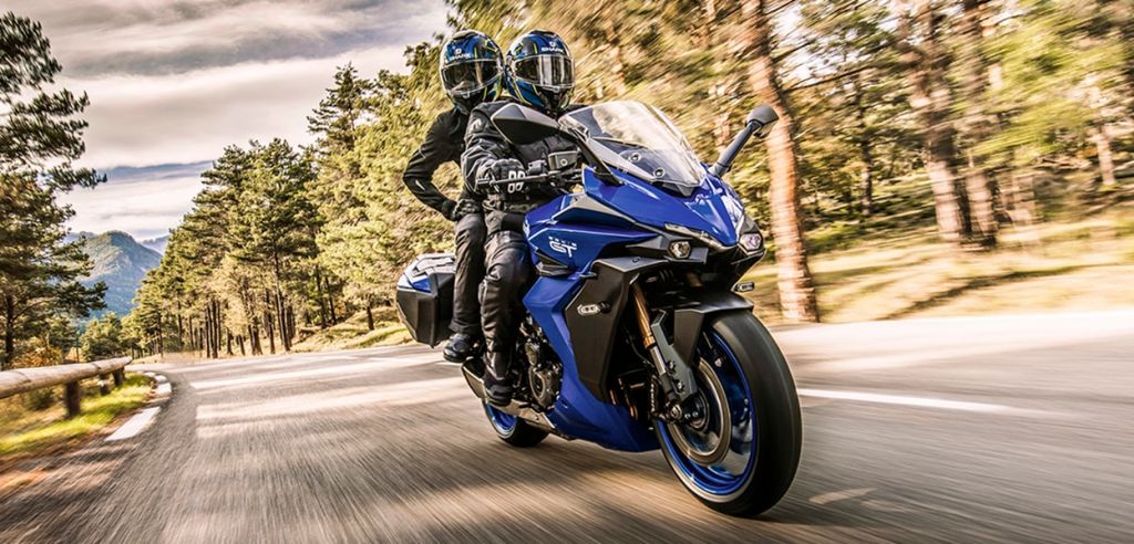 Zwei Motorradfahrer auf einer blauen Suzuki GSX-S1000GT fahren dynamisch durch eine kurvige Bergstraße im Wald.