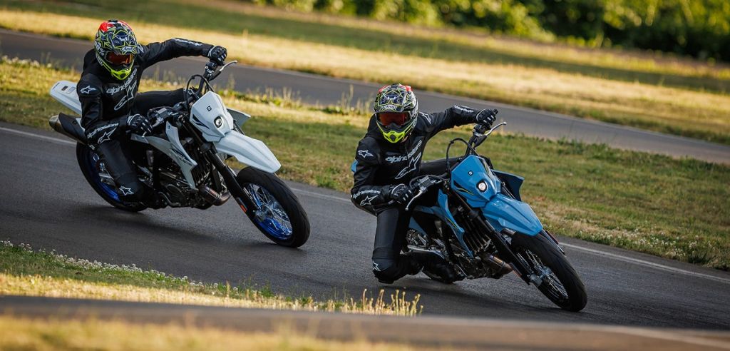 Zwei Fahrer auf sportlichen Dual-Purpose-Motorrädern (weiß und blau) legen sich auf einer Rennstrecke tief in die Kurve.