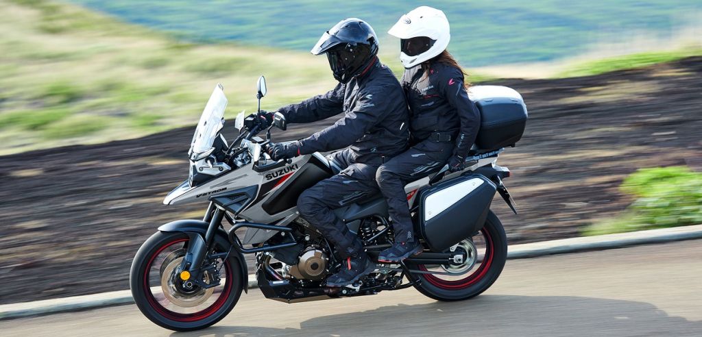 Fahrer und Beifahrerin auf einer grauen Suzuki V-Strom 1050 mit Koffern cruisen über eine Landstraße in weiter, grüner Landschaft.