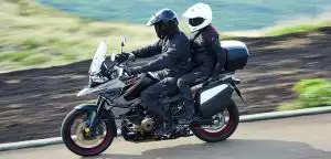 Fahrer und Beifahrerin auf einer grauen Suzuki V-Strom 1050 mit Koffern cruisen über eine Landstraße in weiter, grüner Landschaft.