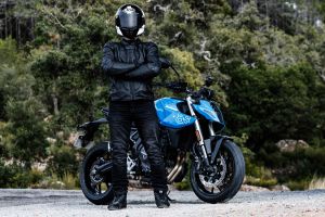 Motorradfahrer in voller Ledermontur und Integralhelm steht mit verschränkten Armen vor einer blauen Suzuki GSX-8S in der Natur