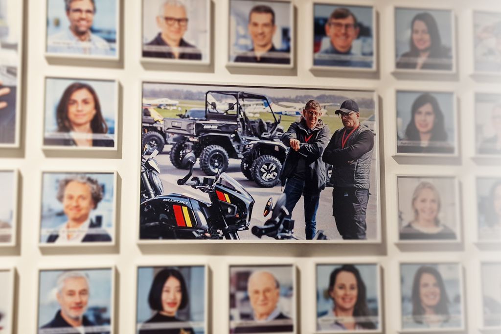 Foto einer Bilderwand mit Red Dot Award Jury: Zwei Männer stehen im Freien vor einer Suzuki GSX-8TT und einem Geländefahrzeug auf einem Flugfeld.
