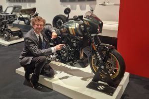 Lead Designer Arthur Vidal im Anzug kniet im Red Dot Museum neben einer dunkelgrün-goldenen Suzuki GSX-8TT auf einem Podest und zeigt auf das Motorrad.