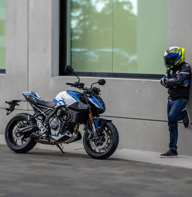 Seitenansicht der Suzuki GSX-8S Special Edition auf dem Seitenständer neben einem an die Fassade gelehnten Motorradfahrer mit Helm.