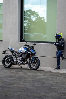 Seitenansicht der Suzuki GSX-8S Special Edition auf dem Seitenständer neben einem an die Fassade gelehnten Motorradfahrer mit Helm.