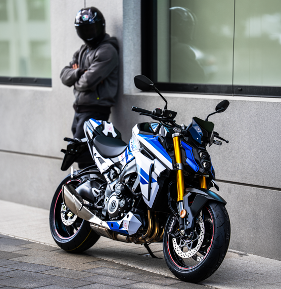 Schrägansicht einer weiß-blauen Suzuki GSX-S1000 Special Edition vor einem modernen Gebäude mit einem Fahrer im Hintergrund.