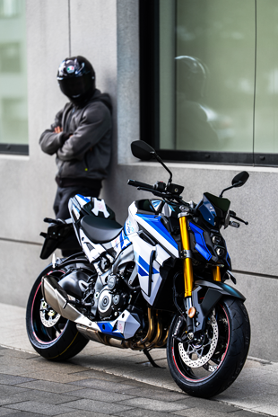 Schrägansicht einer weiß-blauen Suzuki GSX-S1000 Special Edition vor einem modernen Gebäude mit einem Fahrer im Hintergrund.