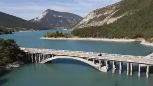 Brücke über türkisfarbenen Bergsee. Berge und Waldlandschaft im Hintergrund. Motorrad fährt über die Brücke.