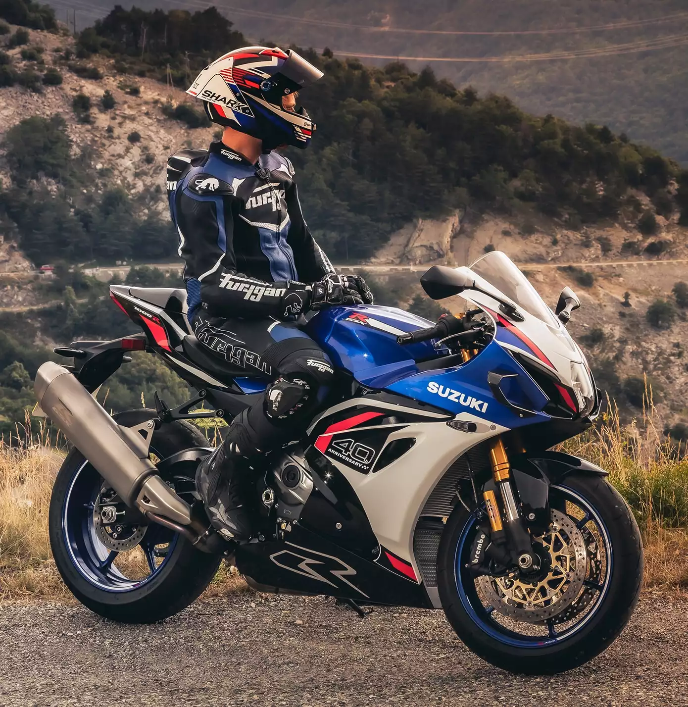 Suzuki GSX-R1000R in Weiß-Blau-Rot mit Fahrer in Rennkleidung, geparkt vor bergiger Landschaft.
