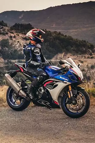 Suzuki GSX-R1000R in weiß-blau-rot mit Fahrer in Rennkleidung, geparkt vor bergiger Landschaft