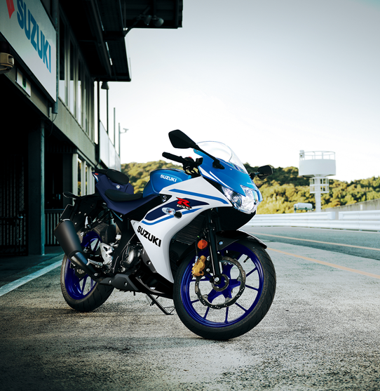 Seitenprofil einer weiß-blauen Suzuki GSX-R125, die auf einem Seitenständer im Fahrerlager einer Rennstrecke steht; im Hintergrund ist das Suzuki-Logo an einem Gebäude zu sehen.