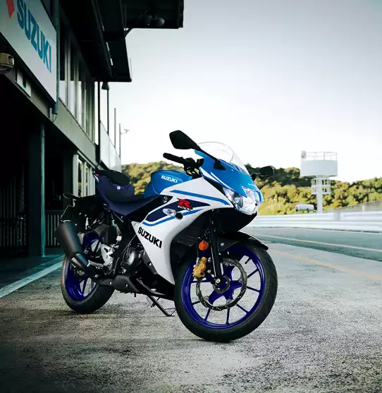 Seitenprofil einer weiß-blauen Suzuki GSX-R125, die auf einem Seitenständer im Fahrerlager einer Rennstrecke steht; im Hintergrund ist das Suzuki-Logo an einem Gebäude zu sehen.